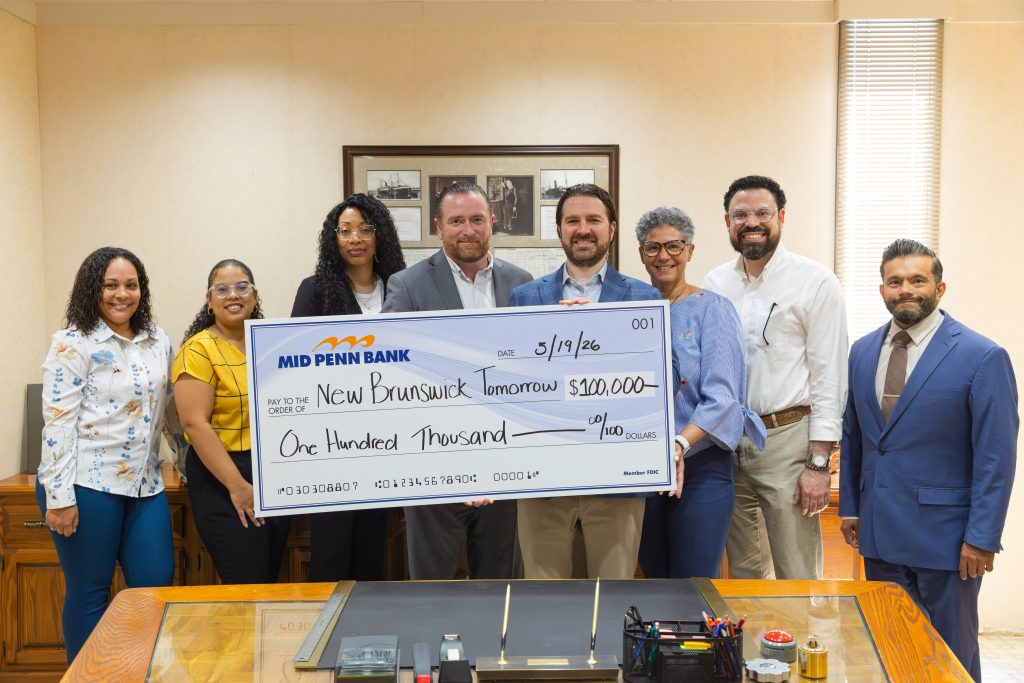Group of professionals standing behind a desk holding a large Mid Penn Bank check made out to “New Brunswick Tomorrow” for $100,000, dated 5/19/26.